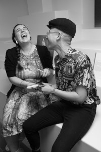 Black and white. A woman with long brown hair throws her head back laughing. A non-binary person next to her is also laughing. They are seated and the woman hands her companion a book.