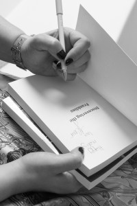 Black and white photo of a woman signing a copy of her book. Close up of hands and book only.