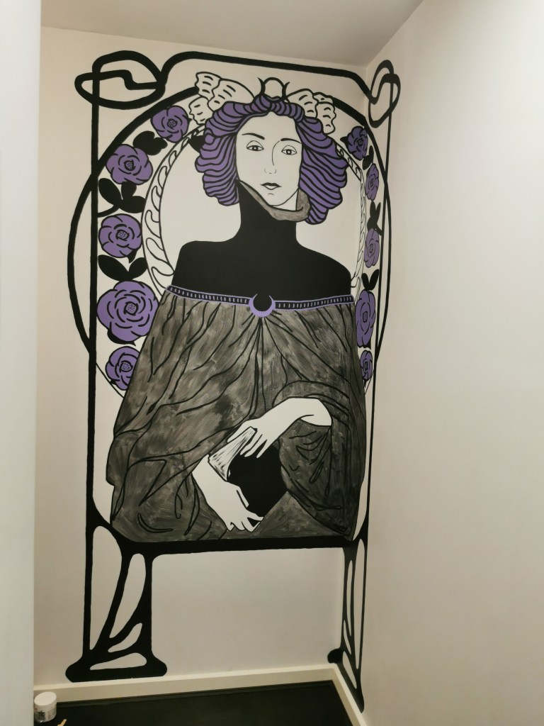 A black, white and purple stylised mural of a woman holding a book. she is surrounded by art nouveau border designs.