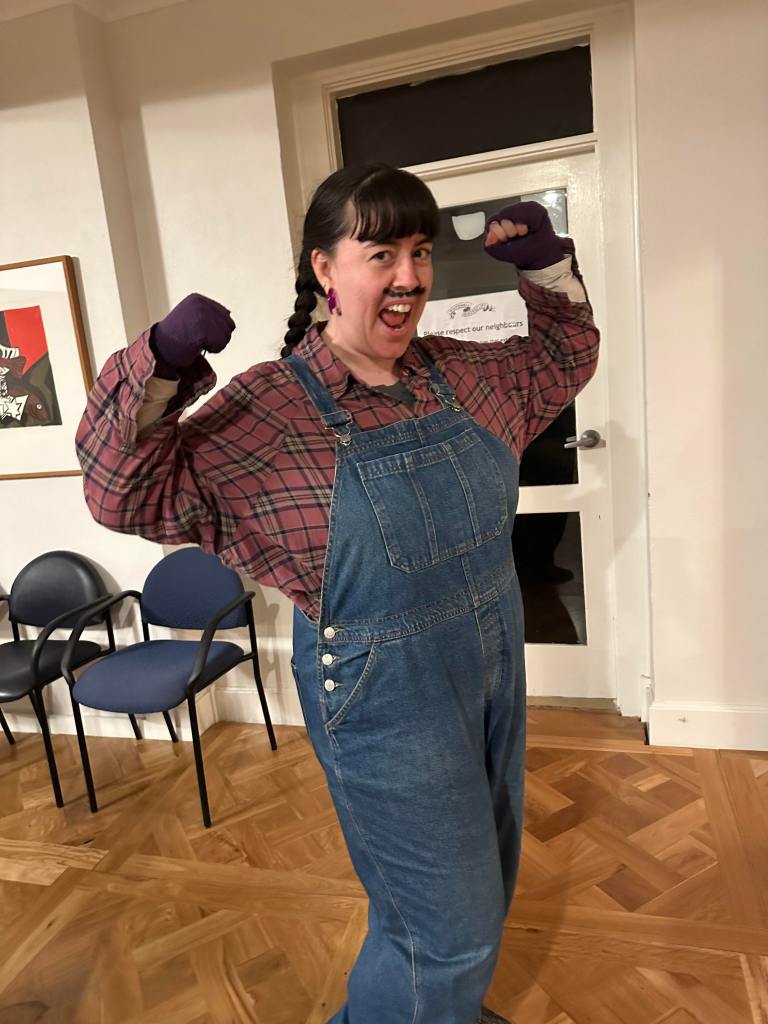 Fleur posing as character Super Mario, she wears a dusty pink check shirt and blue demin overalls. Her arms are up as though flexing her muscles.