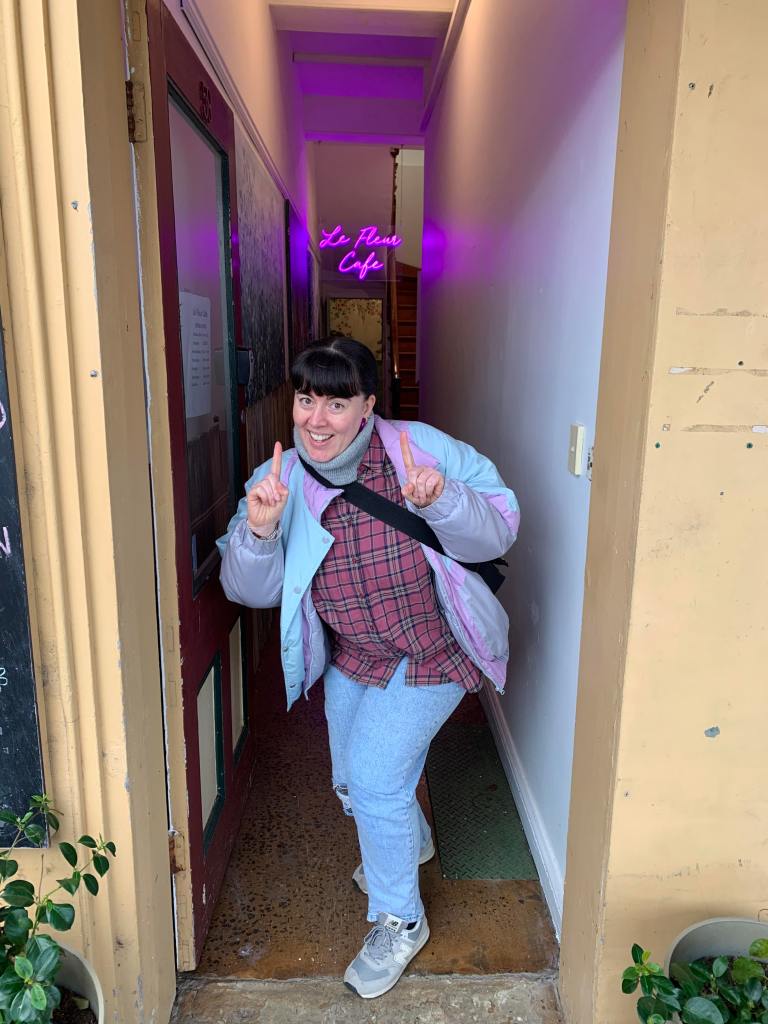 Fleur poses under a sign stating 'Le Fleur Cafe'. She stands in a the entrance of the cafe, with a narrow hallway behind her. She is wearing pale, blue jeans, a check shirt and a pale coloured puffy jacket.