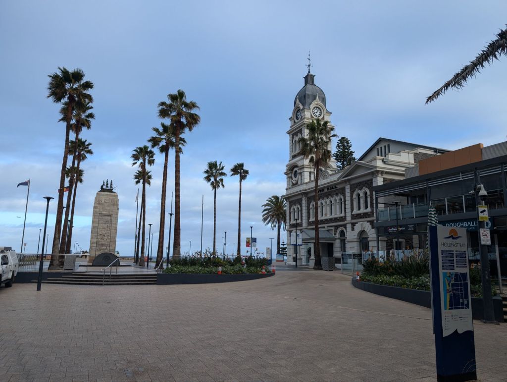 A landscape of Glenelg, SA. The landscape features several palm trees leading to a sandstone war memorial on the left. On the right stands a town hall with a clock tower. The sky is blue, slightly overcast, the foreground shows beige paving.