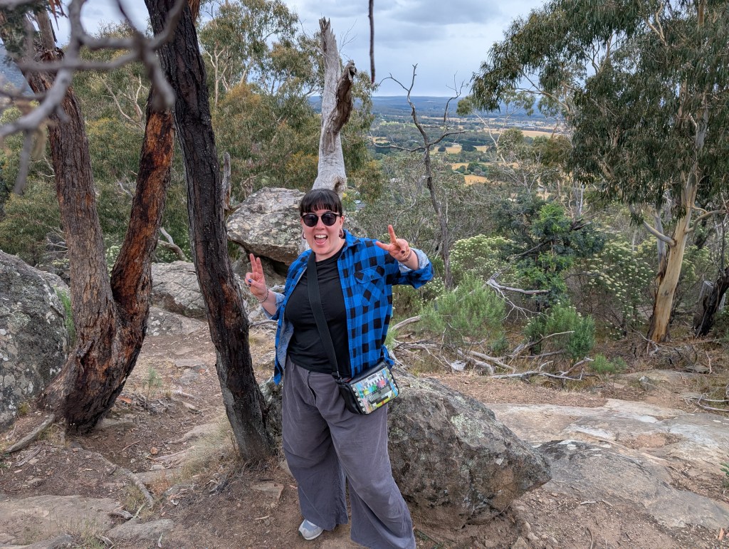 A woman wearing a blue and black checked shirt, black tshirt, and grey pants, stands on a large rock surface in front of a bush vista with trees in the background. She is smiling and making peace signs with her hands.