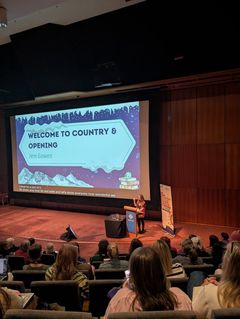 Image of a conference room, with a woman standing at the podium speaking. 