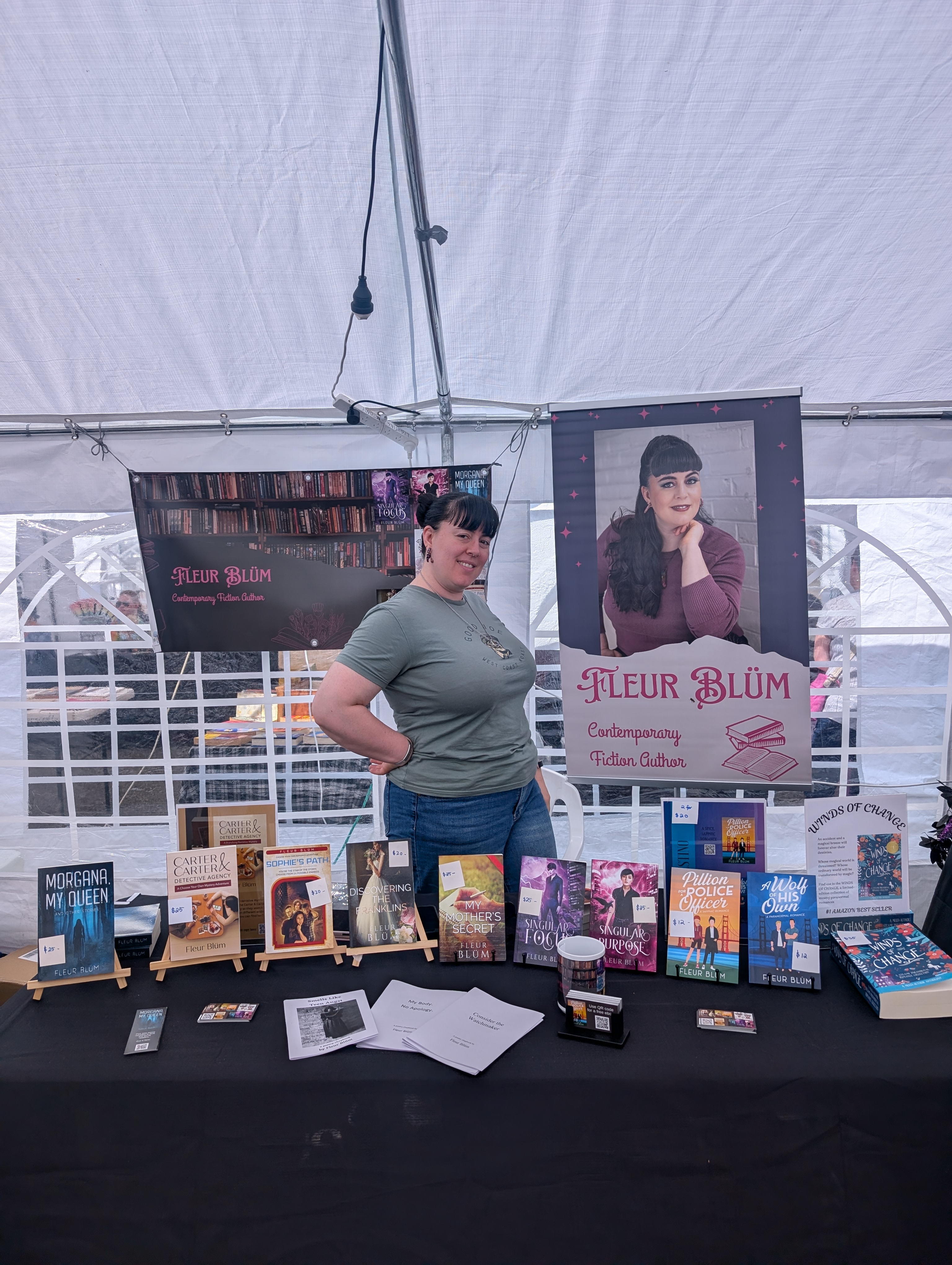 Fleur stands behind a table with a display of her books, in the background is are two banners with Fleur's face and books on them.