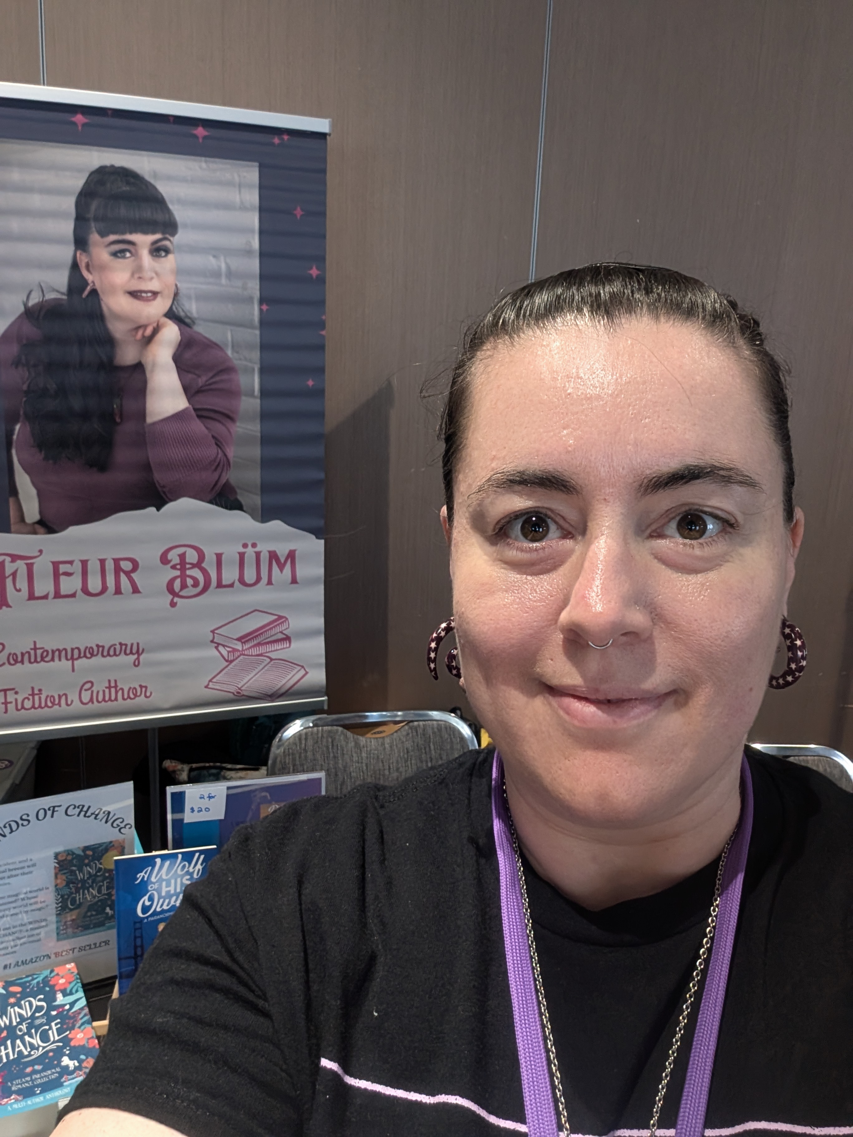 Fleur appears in the foreground, she is smiling and wears a black tshirt. In the background is a banner with Fleur's face on it.