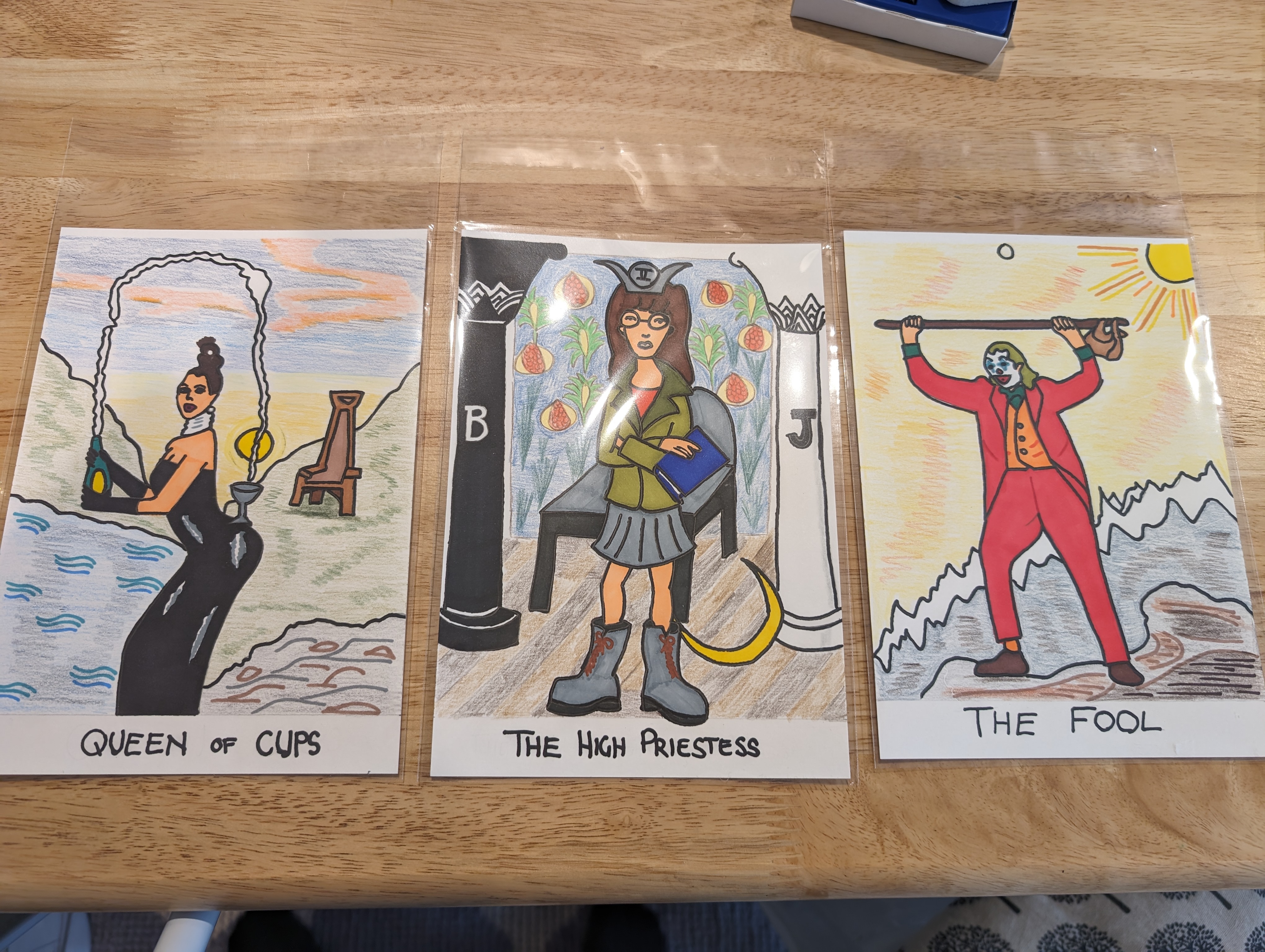 Three cards on a wooden table top, the left drawing features a woman in a black dress holding champagne, and the words 'Queen of Cups', the centre drawing is of cartoon character Daria between two columns, with the words 'The High Priestess', the right hand drawing features a man in a red suit and clown make-up with the words 'The Fool' at the bottom