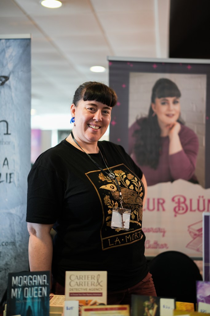 Fleur stands in front of a banner with her author photo. In the foreground we can see a table with books for sale. 